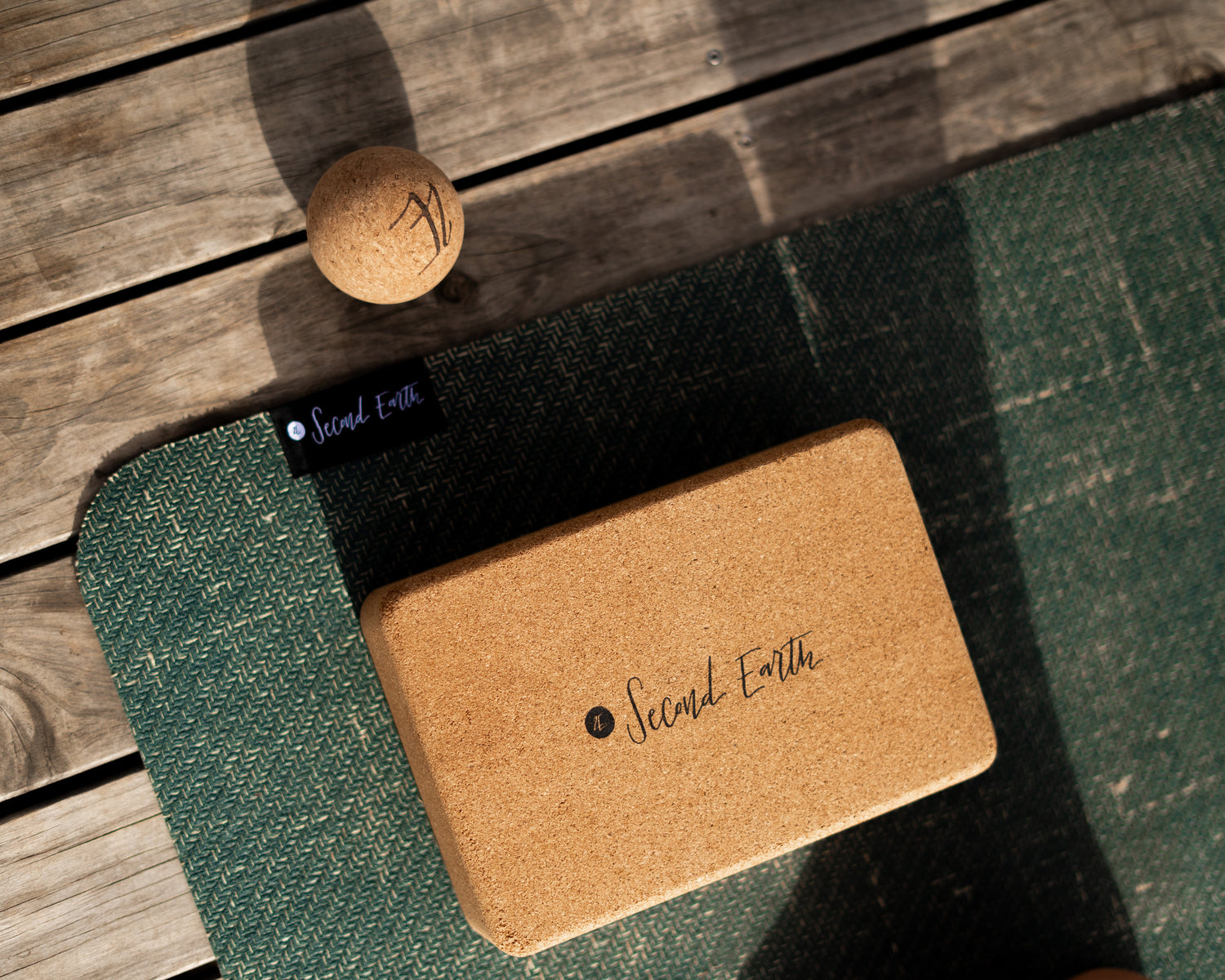A cork yoga block with 'Second Earth' branding resting on a dark green textured Second Earth yoga mat, with a small cork massage ball on a wooden deck background.
