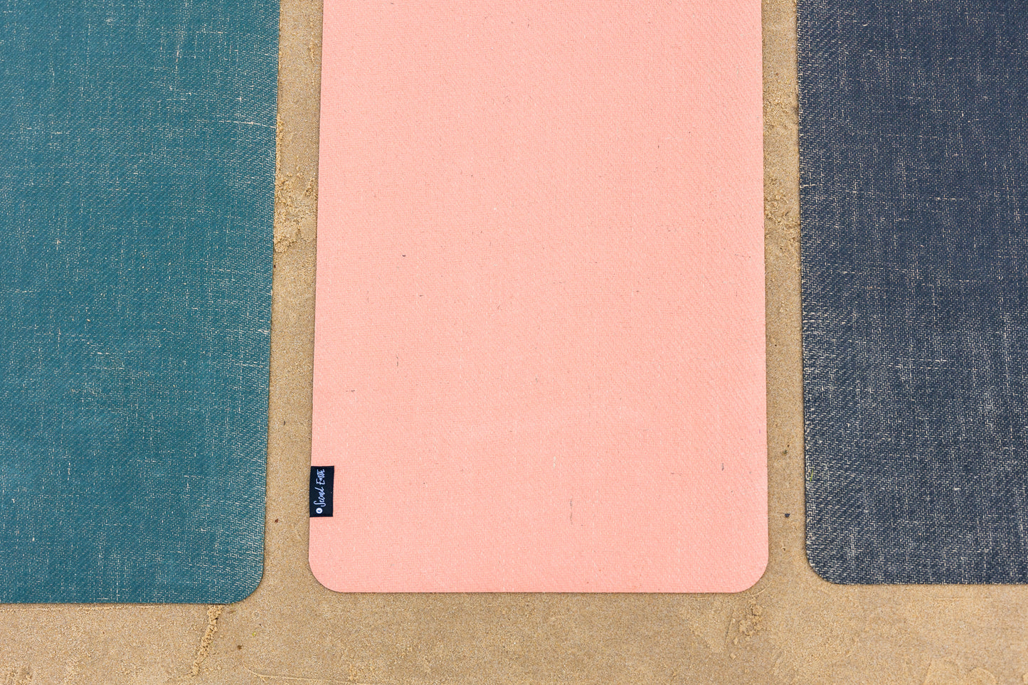 A pink Second Earth yoga mat laid flat on sand surface, flanked by Forest Green and Earth Black textured mats on both sides. 
