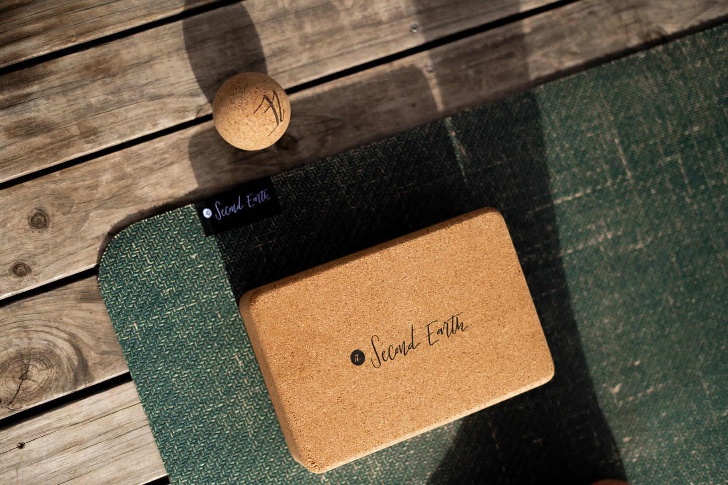 A cork yoga block with 'Second Earth' branding resting on a dark green second earth Connected yoga mat, with a small cork massage ball on a wooden deck background.